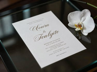 Overhead composition of an elegant event invitation displayed on a dark glass surface — intricate typographic layout glowing softly on screen, a single white orchid bloom placed at the frame's edge, moody studio lighting with turquoise reflection