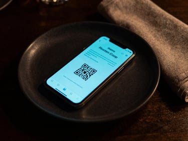 Extreme close-up overhead of a fine-dining table in near-darkness — a single smartphone glowing with a QR-linked menu interface, turquoise light spilling across matte black porcelain and a folded linen napkin, dramatic moody ambient light, no diners visible