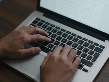 Close overhead view of hands on a laptop keyboard, a website wireframe visible on screen, cool overhead office light, tight crop showing just the keyboard and screen edge