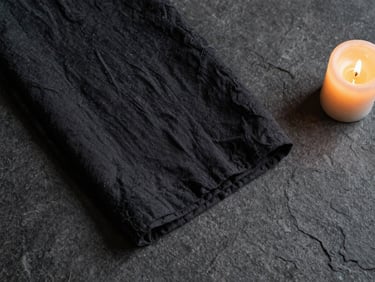 Overhead flat-lay of folded dark linen fabric on a matte stone surface, single candle flame at the corner, warm pooling light, minimal and textural, art-directed editorial