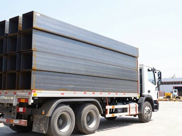 A heavy-duty delivery truck loaded with structural steel beams parked outside a construction site, bright daylight, 35mm lens