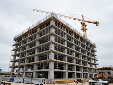 Wide shot of a mid-rise commercial construction site exterior, scaffolding on a concrete frame building, workers on upper floors, construction crane visible against overcast daylight sky, materials staged at ground level