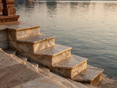 Close-up of carved marble temple steps descending into Udaipur's lake water at golden hour, faint ripple on the water surface, warm reflected light on stone, no people, texture and scale