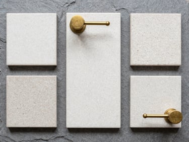 Top-down flat lay of tactile terrazzo tile samples and unlacquered brass fixtures on a neutral slate grey background, bright diffused daylight, sharp focus.
