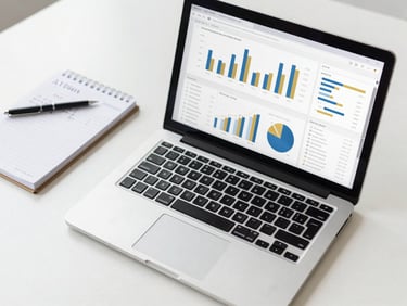 Overhead shot of an analytics dashboard on a laptop with charts and graphs, next to a notepad with handwritten notes, in a clean, minimalist workspace.