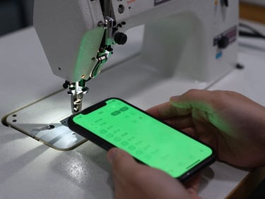 Tight macro shot of a worker's hand tapping a smartphone screen next to an active sewing machine, vibrant green interface, high contrast workshop shadows.