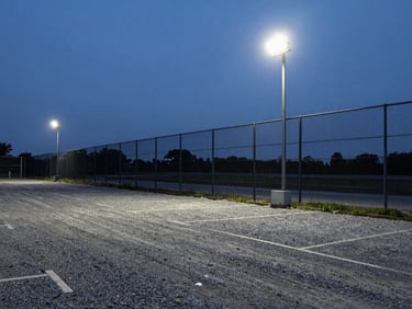Wide angle view of a gravel parking lot lined with high security fencing, bright security floodlight mounted on a pole, evening sky.