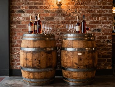 Two bourbon barrels standing side by side as a cocktail-hour bar station inside a reception hall, warm amber interior light, bottles and glassware resting on top, exposed brick wall behind them