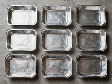 Overhead flat-lay of folded aluminum trays and foil sheets in multiple dimensions arranged in a grid pattern on a concrete surface, cool neutral studio lighting, metallic surface texture sharp and detailed