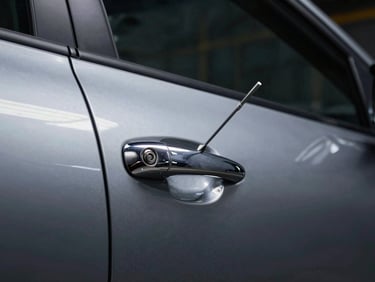 Wide shot of a car door lock cylinder viewed from outside the vehicle, a locksmith pick tool partially inserted, bright workshop fluorescent light hitting the chrome door handle, dark night background