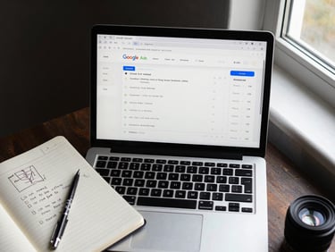 Overhead desktop view of a laptop screen showing a Google Ads campaign dashboard with live click and conversion data, notebook with handwritten funnel notes beside the keyboard, clean window light from the right, high contrast, no faces