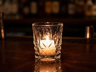 Moody editorial food photography of a dimly lit bar counter, a single heavy crystal glass with condensation catching warm amber candlelight glow, extreme shallow depth of field, 35mm
