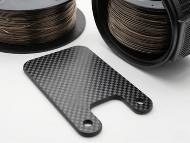 Overhead flatlay of carbon-fibre composite printed bracket beside chopped-fibre filament spool on white surface, bright studio light revealing woven texture and layer precision, tight crop