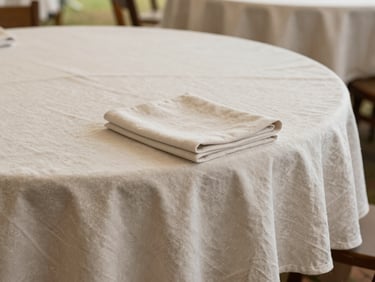 Close detail of neatly pressed linen tablecloths laid over round tables inside a tent, natural daylight from above, fabric folds visible, neutral tones