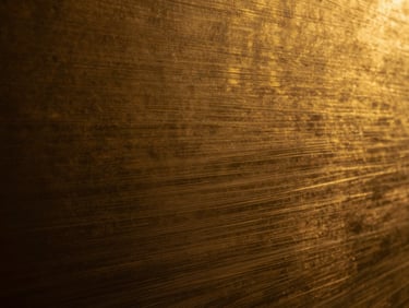 Extreme close-up of hand-brushed brass texture, dramatic low-key lighting showing fine linear scratches, golden-hour warmth.