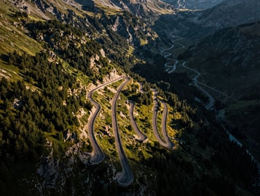 Aerial view of the Stelvio Pass road, showing numerous switchbacks, under dramatic high-contrast light, with the road appearing like a ribbon through rugged mountains.