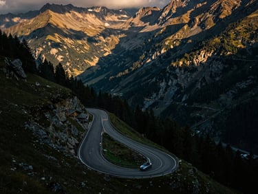 Winding road of Stelvio Pass, viewed from above, with a convertible car small in the frame, surrounded by vast, rugged mountains, dramatic shadows, moody dawn light, wide-angle, cinematic.