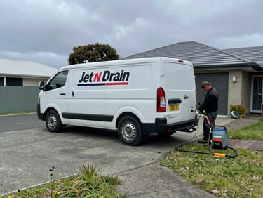 Wide shot of Jet n Drain van parked on a residential driveway, technician working at the rear with jetting equipment laid out, overcast Taranaki daylight, practical documentary framing
