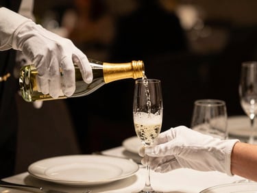 Tight close-up of a white-gloved server's hands pouring champagne into a crystal flute at an intimate private dinner, warm golden ambient light, dark background, no faces