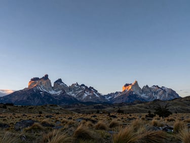 Wide panoramic shot of a Patagonian landscape at dusk, granite peaks in the far right third, open steppe grassland in the foreground catching the last light, vast sky above — anticipation of scale, no people