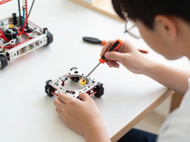 A student assembling a physical robotics kit on a wooden workbench, holding a small metal chassis and screwdriver, bright natural light