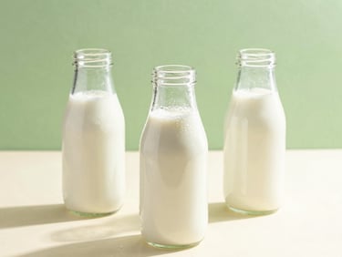 A high-key, bright morning sunlight photograph of condensation-rimmed glass milk bottles sitting on a clean cream surface, 50mm lens, fresh green accents in the background.