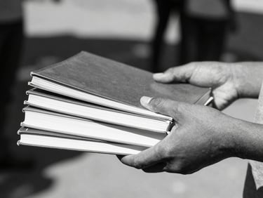 Close-up of hands passing a stack of new schoolbooks in bright South African sunlight, crisp black and white tones, shallow depth of field.