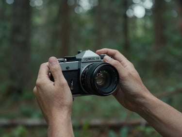 moody cinematic film photography, close-up of hands adjusting a classic 35mm film camera lens, natural overcast daylight, deep forest background