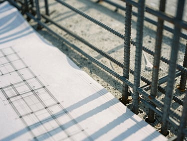 Tight macro shot of architectural blueprints overlaying raw concrete and steel reinforcement bars, crisp morning daylight, cool-toned, architectural film photography.