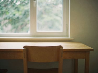 A quiet, minimalist study room with a single chair, wooden table, and large window showing green foliage, warm editorial photography, 35mm