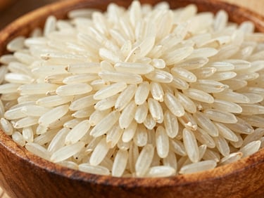 Close-up of raw, polished long-grain Basmati rice grains in a rustic wooden bowl, bright daylight.