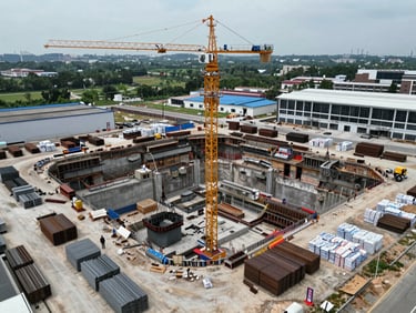 Aerial wide shot of a large commercial mixed-use development site, tower crane centered over a deep excavation with concrete pour in progress, organized material staging areas with stacked steel sections and palletized cement bags visible at the perimeter, overcast diffused daylight