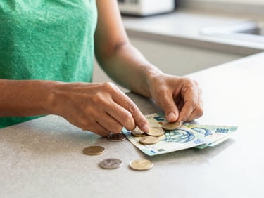 Close-up of a Brazilian woman's hands carefully counting coins and small bills on a kitchen counter, bright natural daylight, soft shadows, warm tones, clean editorial photography, soft green and blue accents, realistic domestic setting, 35mm