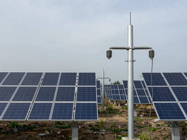 Wide-angle ground-level view of a utility-scale ground-mount solar array under overcast daylight, rows of panels extending to the horizon, a high-voltage riser conduit in the foreground showing electrical integration precision, no people