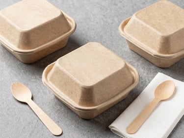 Crisp flat-lay of eco-friendly brown paper food containers, wooden cutlery, and clean white napkins on a neutral slate background, studio lighting