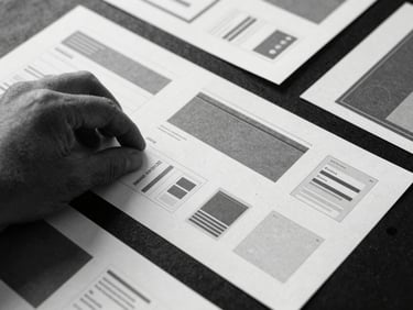 Stark brutalist flat lay photography, flat scanner-bed lighting, high-contrast monochrome with raw textures, sharp macro focus of a hand arranging modular paper UI wireframes on a dark surface.