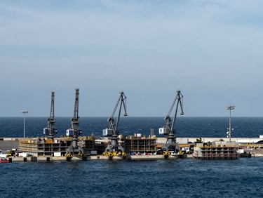 Wide-angle shot of a modern port under construction, showing heavy machinery, concrete structures, and the sea, overcast coastal daylight, deep blues and steel grays, high contrast.