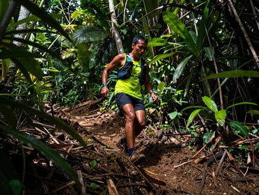 Trail runner ascending a steep, muddy hill, dense tropical vegetation, sweat and determination on face, low-angle.