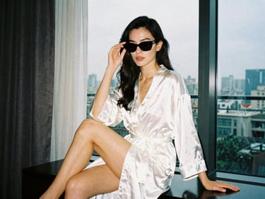 A glamorous girl in her luxury hotel room wearing a white silk robe and black sunglasses, sitting down showing her long legs, dramatic window lighting, 35mm film style