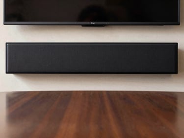 Direct front-on studio shot of a home soundbar, slim black profile, mounted below a screen in a meeting room setting, neutral background, even studio lighting showing grille texture