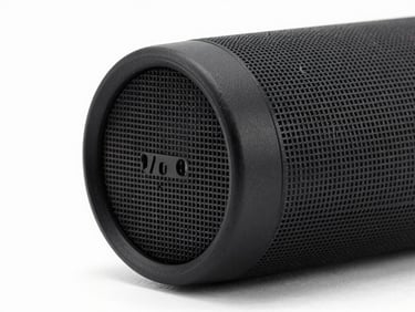 Studio close-up of a portable Bluetooth speaker, cylindrical form, black matte finish, front grille detail visible, clean white background, controlled strobe lighting