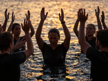 Dramatic outdoor baptism service at dusk with hands raised in worship, warm golden firelight reflections on water, 35mm