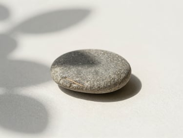 Close-up of a smooth, grey pebble on a soft, off-white surface, with a subtle shadow of a single leaf falling on it, soft natural daylight.
