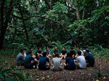 Wide shot of a small group gathered in a circle on a quiet forest floor, diffused overcast daylight, deep green and indigo tones