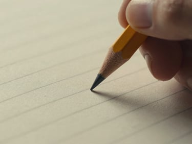Close detail of a pencil tip moving across a ruled notebook page, fingers partially visible, soft warm overcast light, grain visible, intimate documentary framing