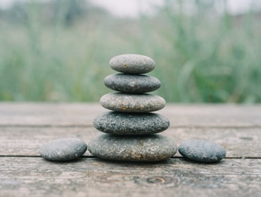 A stack of smooth grey river stones arranged neatly on a weathered wooden plank, soft-focus background of gentle green foliage.