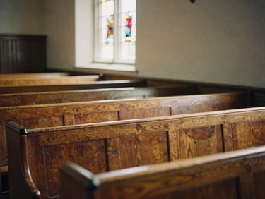 Worn wooden pews inside a quiet sanctuary, soft natural light streaming through a stained glass window, warm historic atmosphere, 35mm film aesthetic