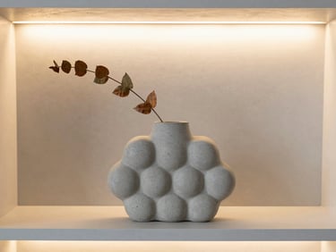 A modern concrete bubble vase with a single dried botanical branch on a light gray shelf, soft warm ambient light