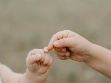 Medium close-up of a baby's hand gently grasping an adult's finger, soft natural daylight, muted earth tones, shallow depth of field, outdoor setting