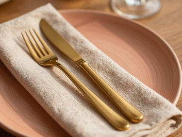 Close-up of a textured ceramic plate with a custom linen napkin and gold cutlery, warm sunlight, shallow depth of field